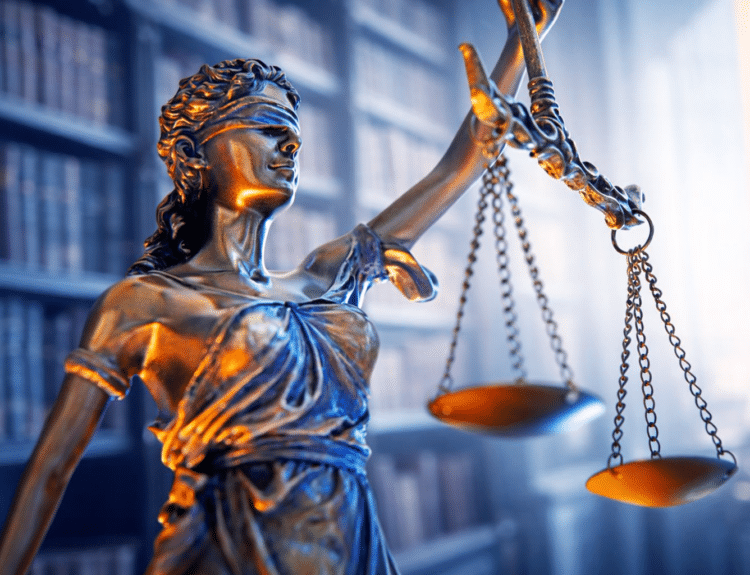 Statue de la Justice aux yeux bandés tenant une balance, éclairée par des lumières bleues et orangées, devant une bibliothèque floue.