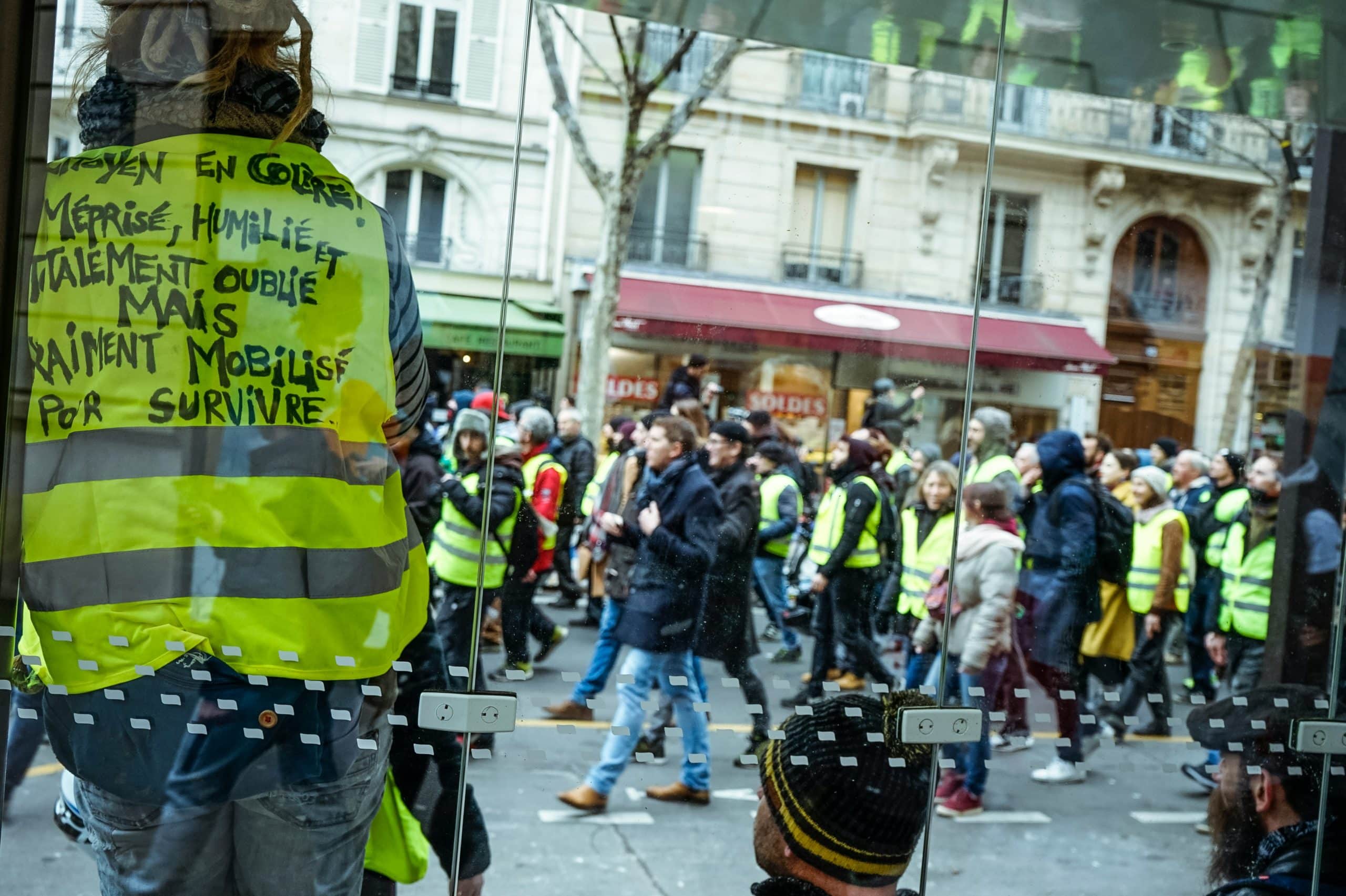 Manifestation de gilets jaunes dans une rue parisienne, avec un slogan écrit sur un gilet au premier plan vu à travers une vitre
